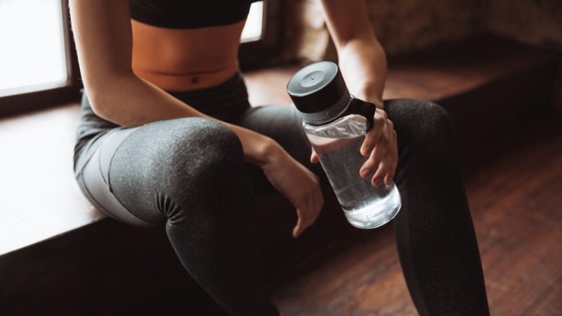 A woman in workout clothes sits on a bench holding a clear water bottle after training