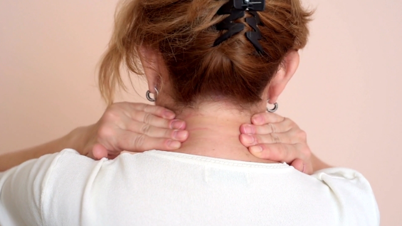 A woman holds the back of her neck with both hands, showing visible discomfort from neck pain