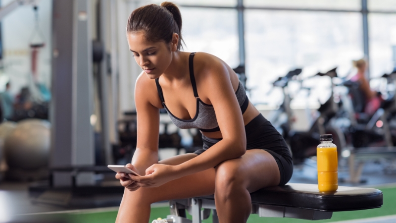 A woman sits on a bench at the gym checking her phone after a workout, with a bottle of juice beside her