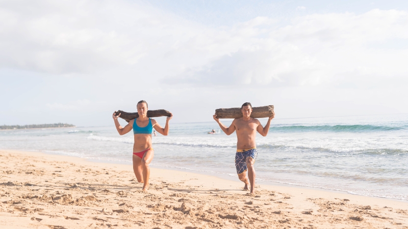 Man and woman exercising on the beach with logs, staying active and healthy while traveling