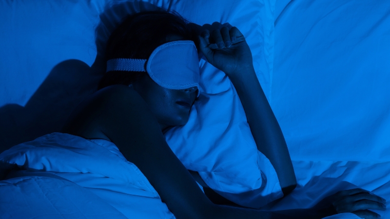 A person sleeps in bed wearing an eye mask, resting under blue lighting that suggests a focus on nighttime recovery