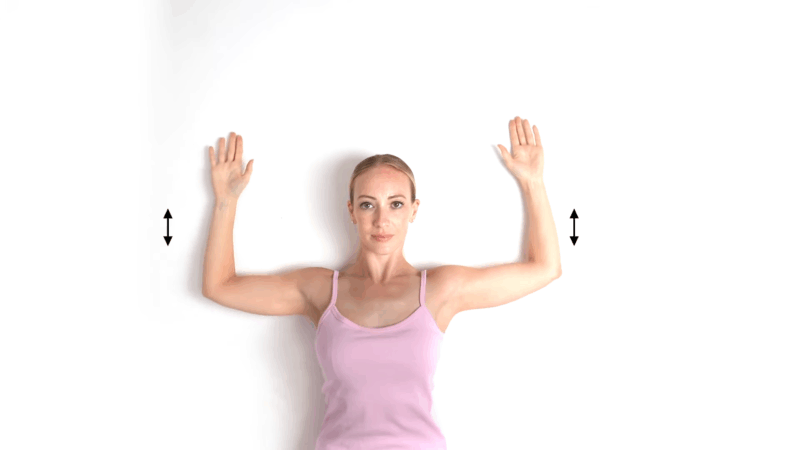 Woman doing wall angels exercise with arms raised against a white wall to improve posture and shoulder alignment