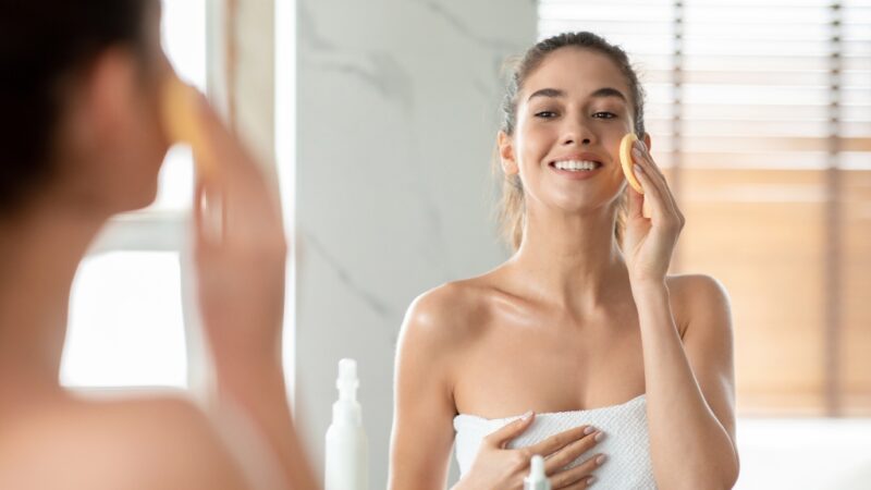 A young woman in a towel smiles while applying skincare to her face with a sponge in a bright bathroom