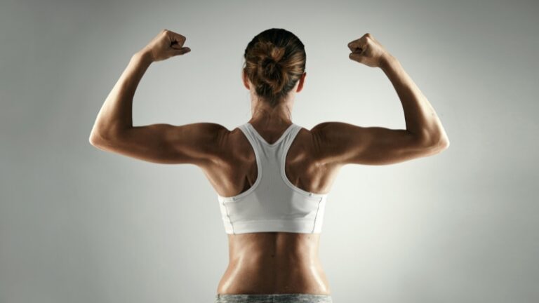 A fit woman in a white sports bra flexes her arms to show strong forearms and defined upper body muscles