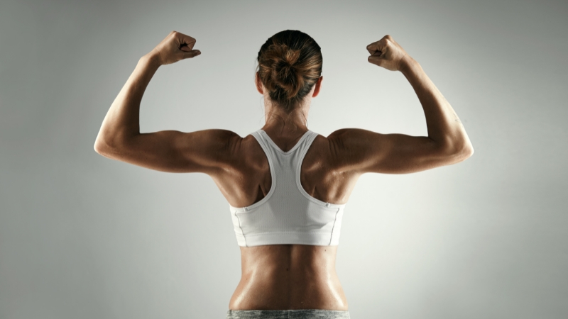 A fit woman in a white sports bra flexes her arms to show strong forearms and defined upper body muscles