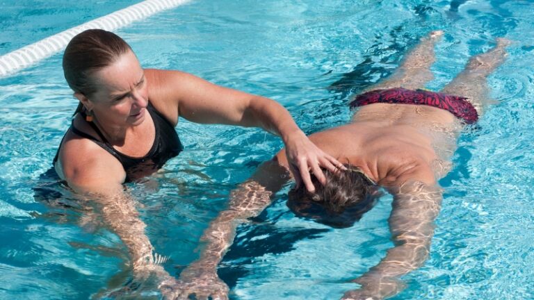 A swimming instructor holds a swimmer’s head and supports their body in the water to practice safe floating techniques
