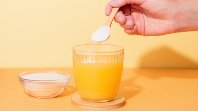 A spoonful of white collagen powder is added to a glass of orange juice on a wooden coaster