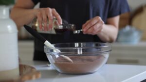 A person pours vanilla extract from a bottle into a measuring spoon over a bowl of mixed dry ingredients on a kitchen counter