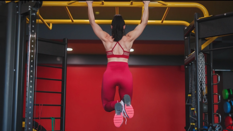 A woman in red workout clothes hangs from a pull-up bar, training grip and building strong forearms
