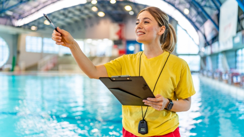 A swimming instructor stands by the pool with a clipboard and whistle, giving directions to students during a lesson