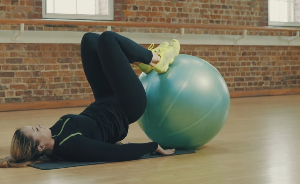 Woman doing hamstring curl with legs on a green stability ball on studio floor