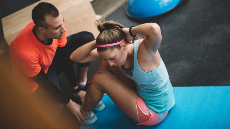 A fitness instructor helps a woman do sit-ups on a mat in the gym