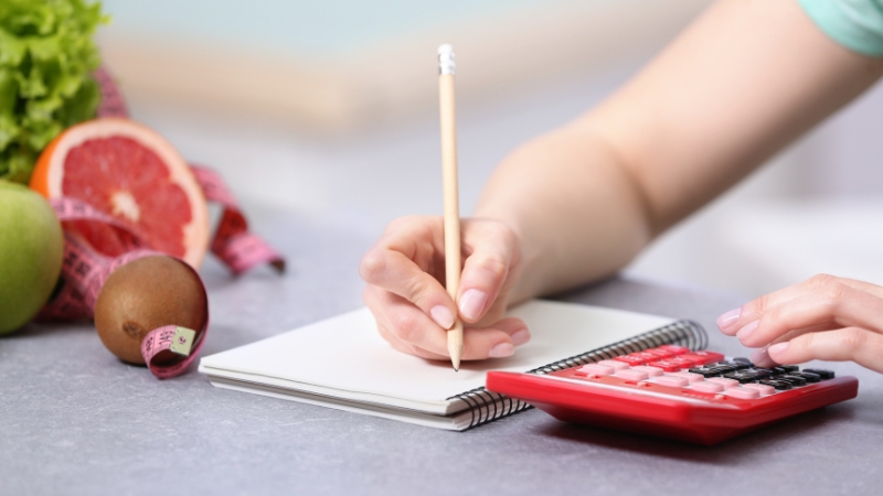 Person using a calculator and notebook to track daily nutrition with fresh fruits nearby
