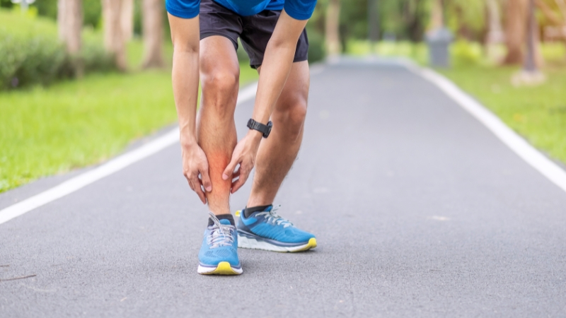 A runner stops on a paved path to hold his lower leg, showing clear shin splints pain during exercise