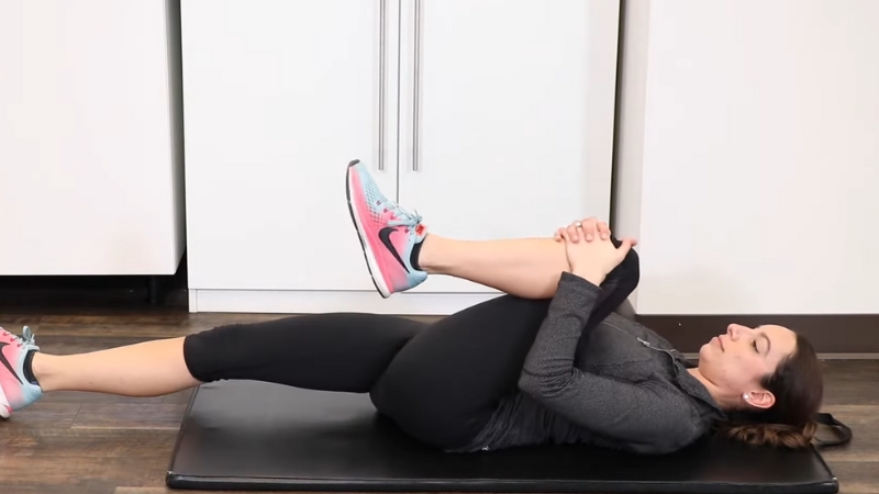 A woman lies on a mat pulling one knee to her chest to stretch her lower back and relieve sciatic pain