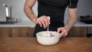 A person in a black shirt is stirring protein powder into a bowl of oatmeal on a wooden counter