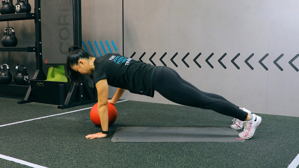 Woman performing push-ups with one hand on a red medicine ball