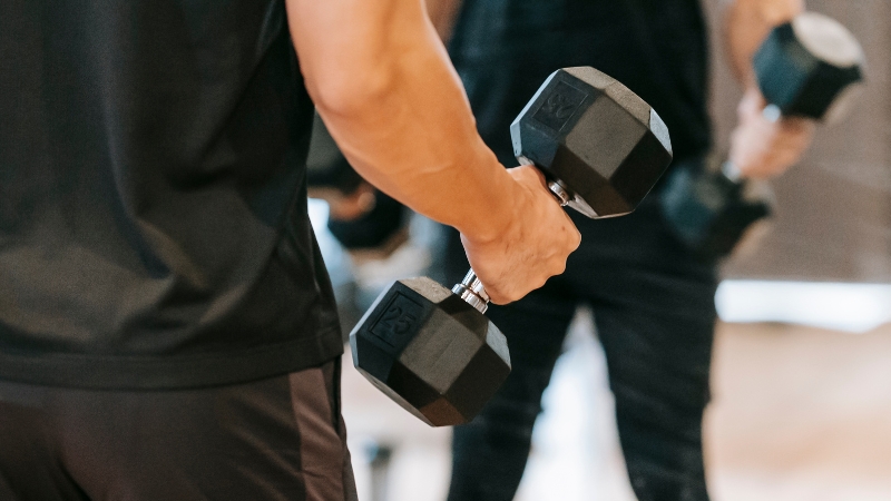 A person lifts dumbbells in front of a large gym mirror that reflects the workout area and adds depth to the space