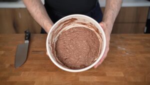 A person is holding a bowl of chocolate protein oatmeal mixture over a wooden counter next to a kitchen knife