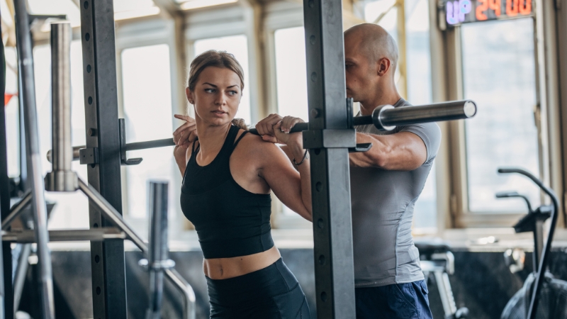 A female client performs squats with a barbell while a male fitness instructor stands behind her, spotting