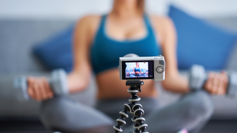 A woman records a fitness video at home with a camera on a tripod while holding dumbbells