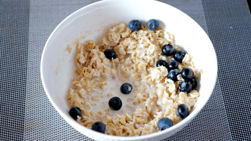A bowl of protein oatmeal topped with fresh blueberries and a splash of milk on a gray placemat