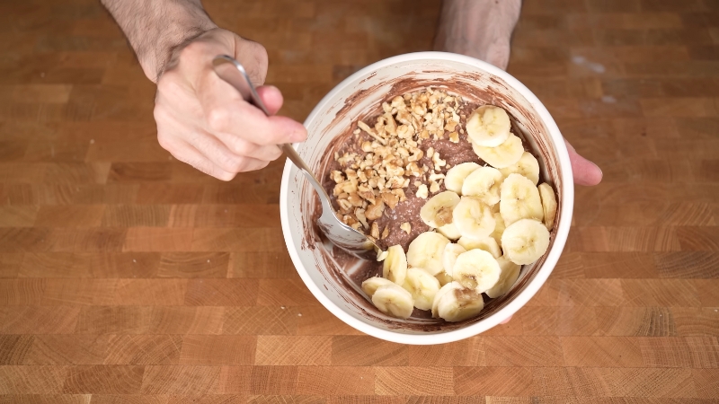 A bowl of chocolate protein oatmeal topped with sliced bananas and chopped walnuts, held in one hand while stirring with a spoon