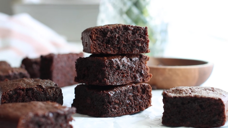 A stack of moist, fudgy high-protein brownie squares with more brownies arranged around them on parchment paper