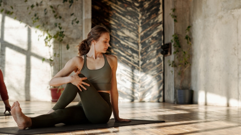A woman practices a seated spinal twist on a yoga mat in a sunlit room