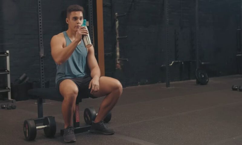 Man in a gym sitting on a bench and drinking from a shaker bottle after a workout.