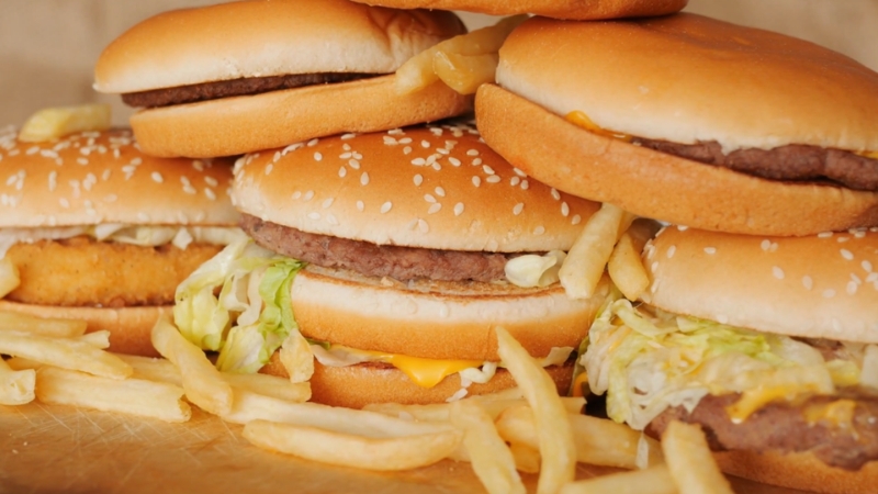 Several fast food burgers and fries are stacked on a wooden surface, showing high-fat, low-nutrient options