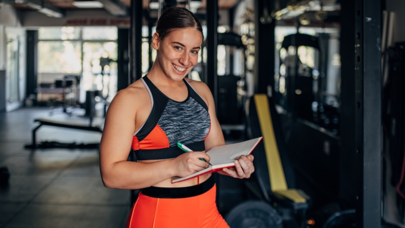 A young woman in workout clothes smiles while holding a notebook in a gym, representing the start of a fitness coaching career