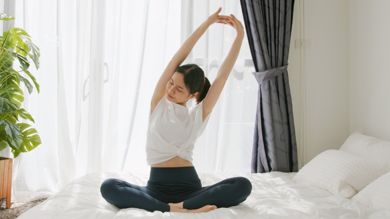 A woman sits cross-legged on her bed, stretching her arms overhead