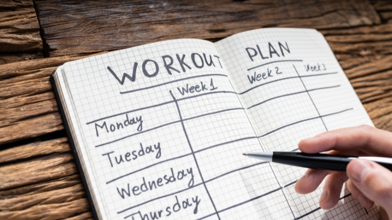 A hand holding a pen over an open notebook with a blank weekly workout plan layout
