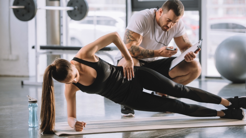 Trainer supervising a woman doing a side plank on a mat at the gym