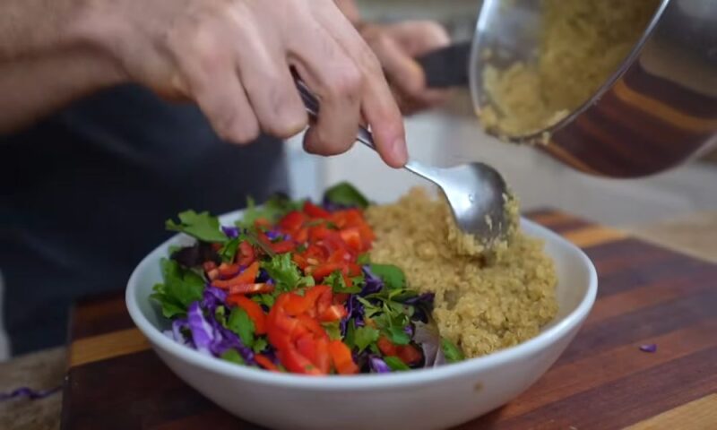 Close-up of quinoa being served into a bowl with fresh vegetables like red peppers, cabbage, and greens.