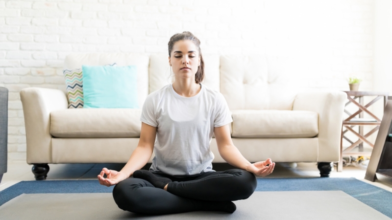 A woman sits cross-legged on a yoga mat with eyes closed, meditating quietly in a bright living room