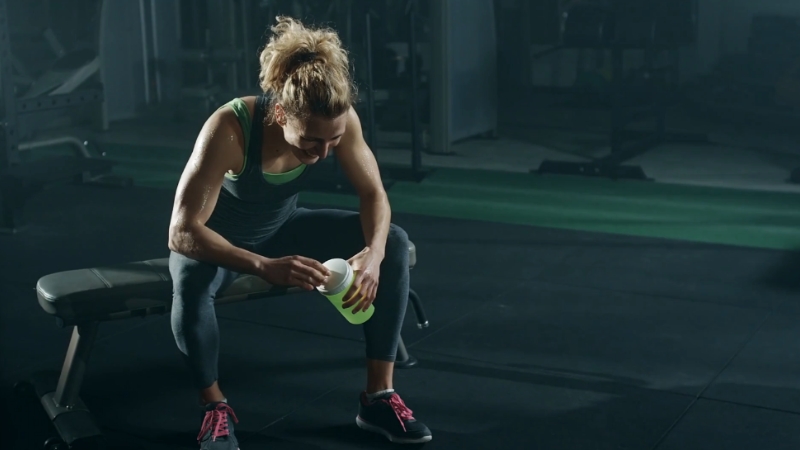 A woman sits on a gym bench preparing her drink after a workout