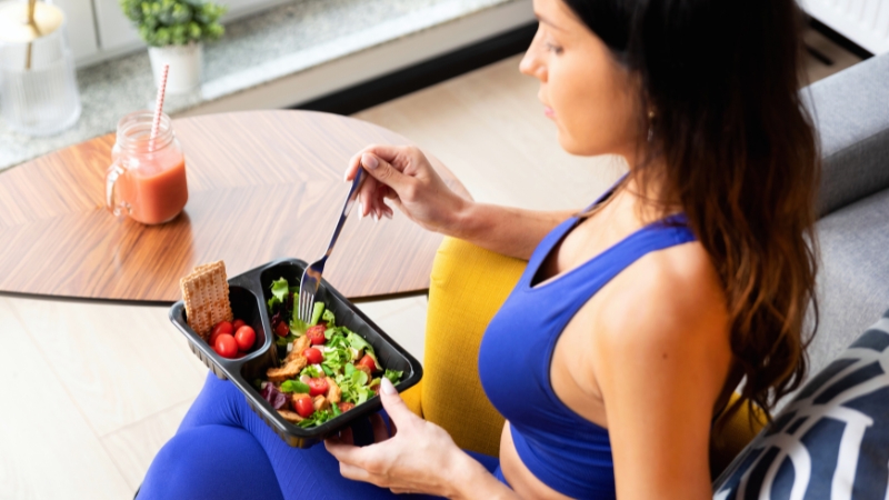 A woman in workout clothes eats a fresh post-workout meal with salad, cherry tomatoes, and crispbread next to a smoothie