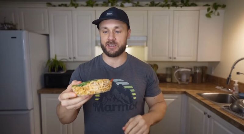 Man in a kitchen holding a bowl of noodles with vegetables.