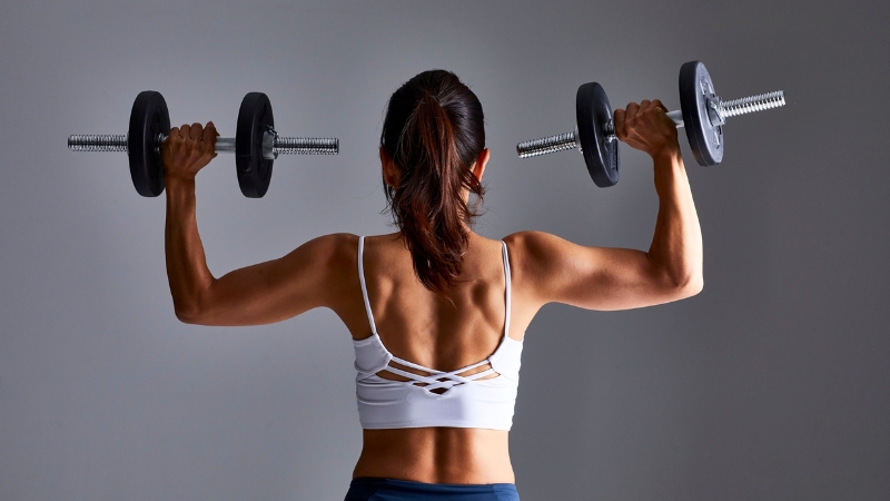 A woman with a ponytail lifts dumbbells behind her head, showing an example of safe hairstyles for workouts
