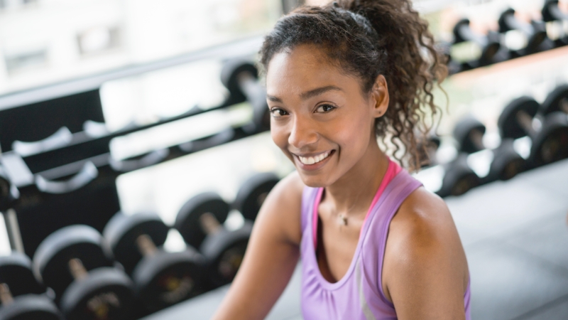 A woman smiling after a workout while sitting near a rack of dumbbells
