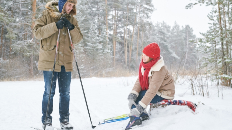 A skier kneels in the snow to check an injury as a partner stands nearby with poles