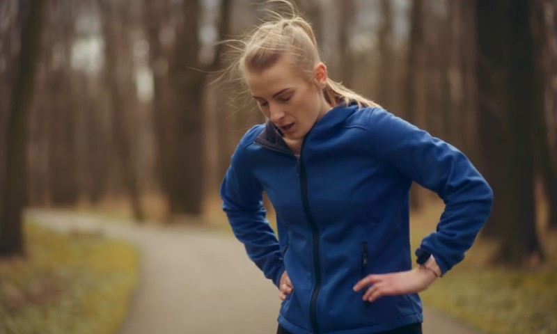 Young woman in a blue jacket pants with hands on hips, appearing tired after running 