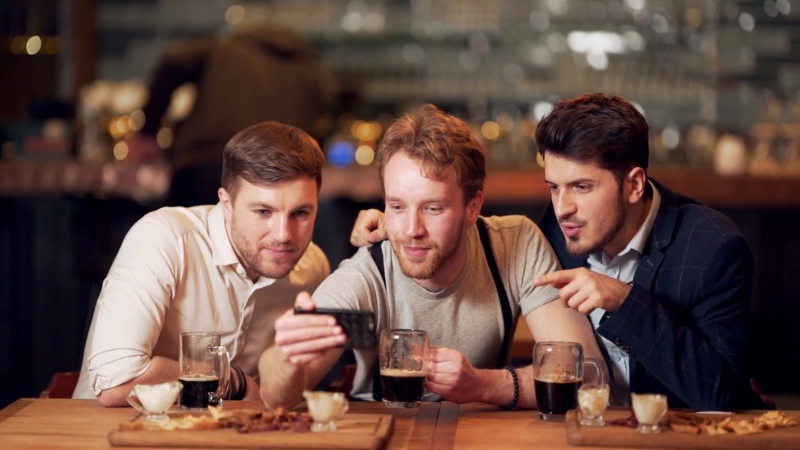 Three friends watch sports highlights on a phone while sitting at a bar
