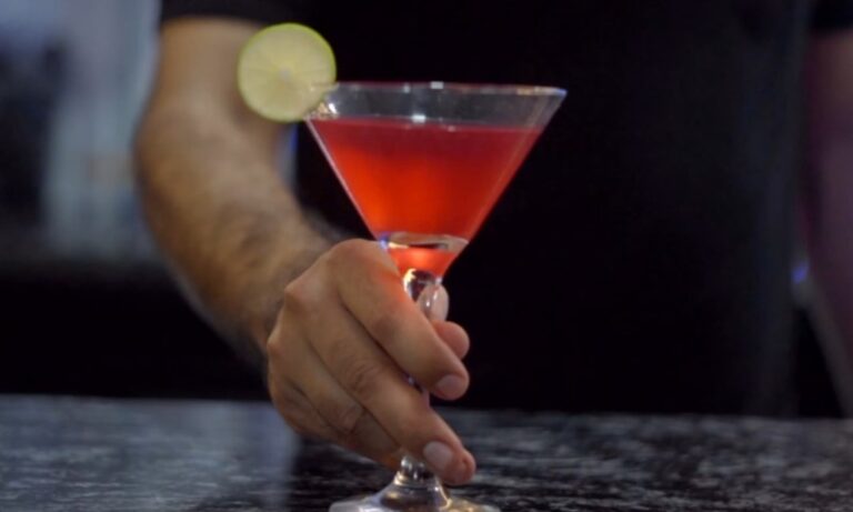 A hand holds a vibrant red cocktail in a martini glass, garnished with a lime slice