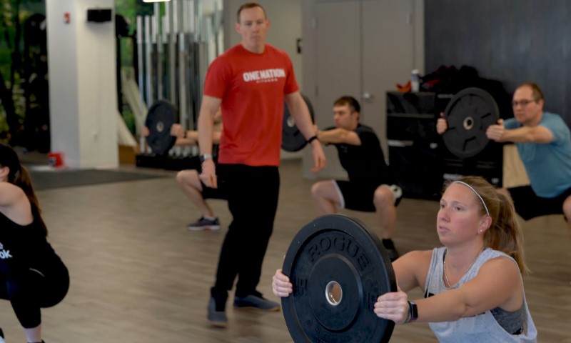 Group fitness class with five people in a gym performing squats, holding weight plates