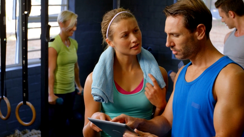 A coach reviews workout data on a tablet with a client inside a gym