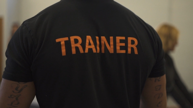 A personal trainer stands with his back to the camera wearing a shirt labeled “Trainer”