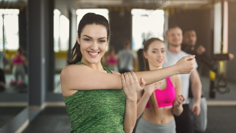 Woman stretches her arm in a gym with others behind her